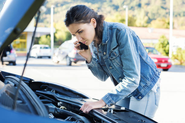A woman holds her cellphone to her ear with one hand while inspecting the inside of her cars hood with the other.